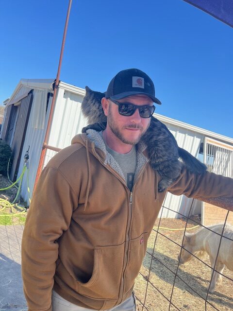 Brian, co-founder of Hidden Pastures, with a cat on his shoulder outdoors on the farm