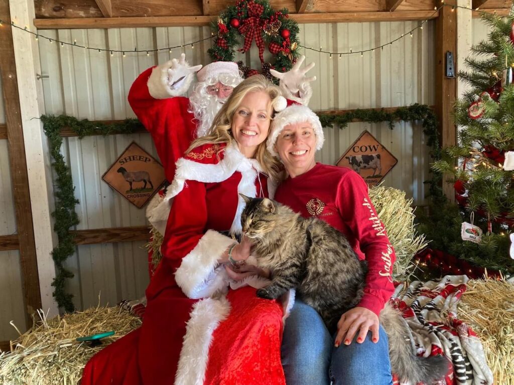 Two people in holiday attire holding a cat in a decorated barn for Holiday Farm Day at Hidden Pastures