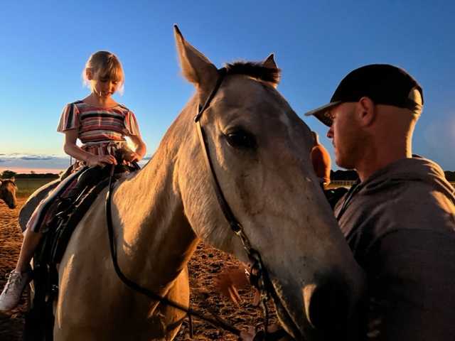 A child riding a horse at sunset while an adult leads and supervises at Hidden Pastures